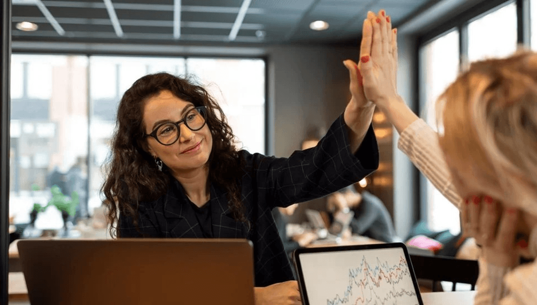 Två kvinnor som gör high five. Den ena kvinnan har mörkt hår, glasögon och ser glad ut. Den andra kvinnan sitter med ryggen mot.